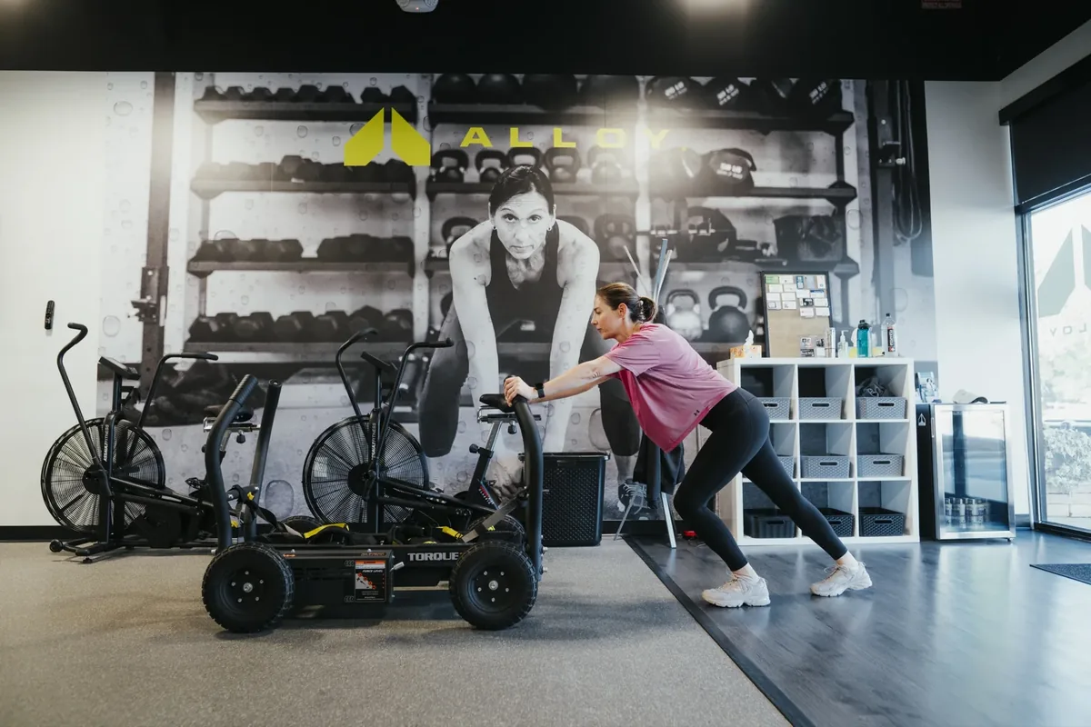 A member in a pink shirt pushes a training sled across the floor at an Alloy Personal Training studio