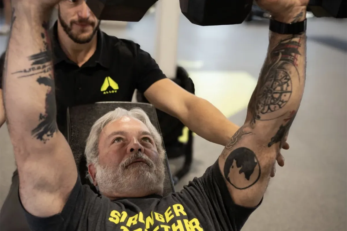 An experienced lifter presses two dumbbells overhead while a coach spots from behind at Alloy Personal Training Middleton in Middleton, WI.