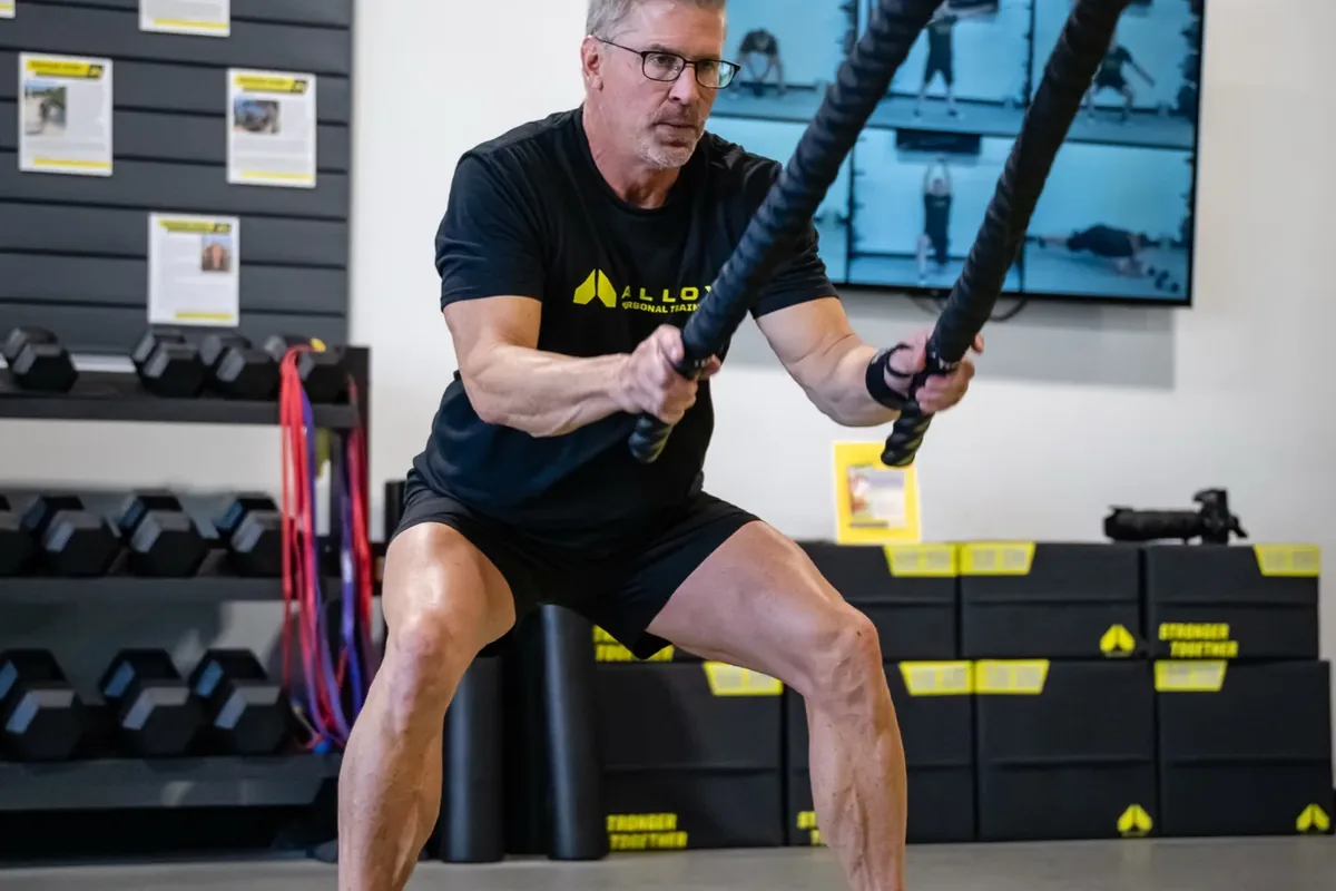 A gray-haired member works battle ropes in a wide stance on the training floor at Alloy Personal Training Middleton in Middleton, WI.