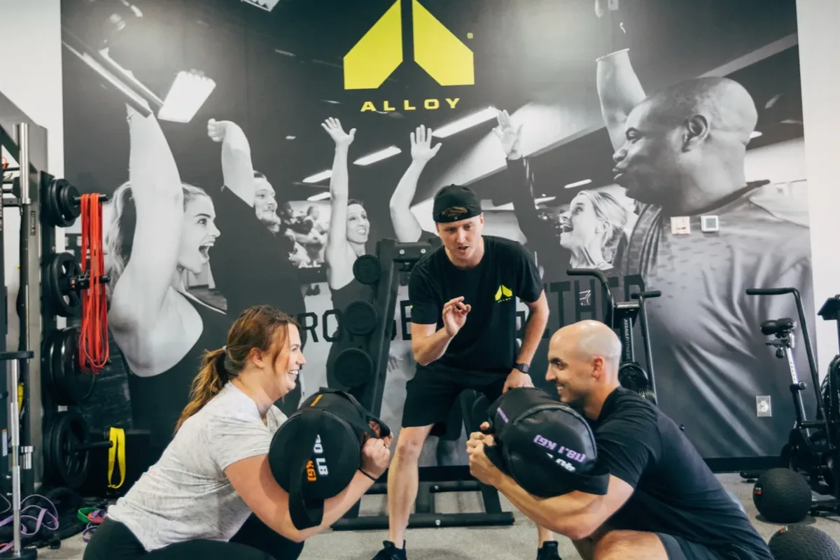 A coach guides a couple squatting face-to-face with sandbags during a small group session at Alloy Personal Training Middleton in Middleton, WI.