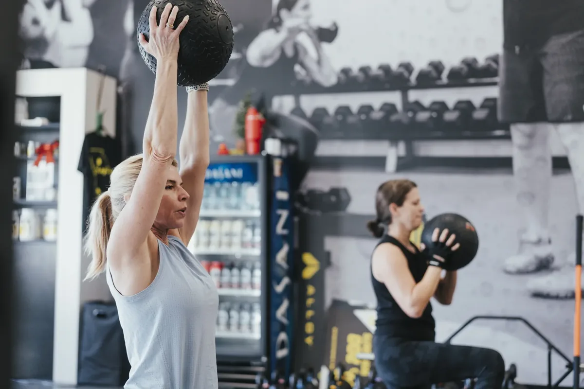 Two members train side by side with slam balls, one pressing hers overhead, at Alloy Personal Training Middleton in Middleton, WI.