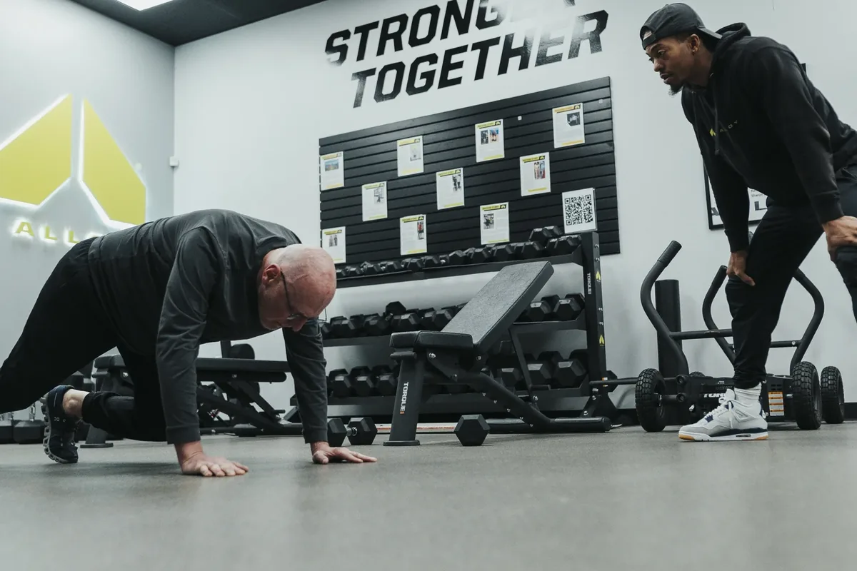 A coach leans in to guide an older member through a push-up during a beginner-friendly session at Alloy Personal Training Middleton in Middleton, WI.