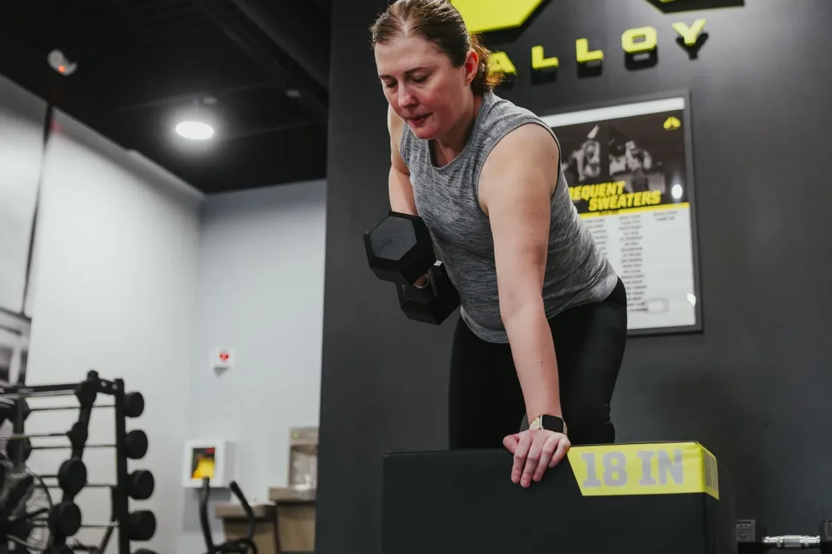 A member performs a single-arm dumbbell row on her own, braced on a plyo box, at Alloy Personal Training Middleton in Middleton, WI.
