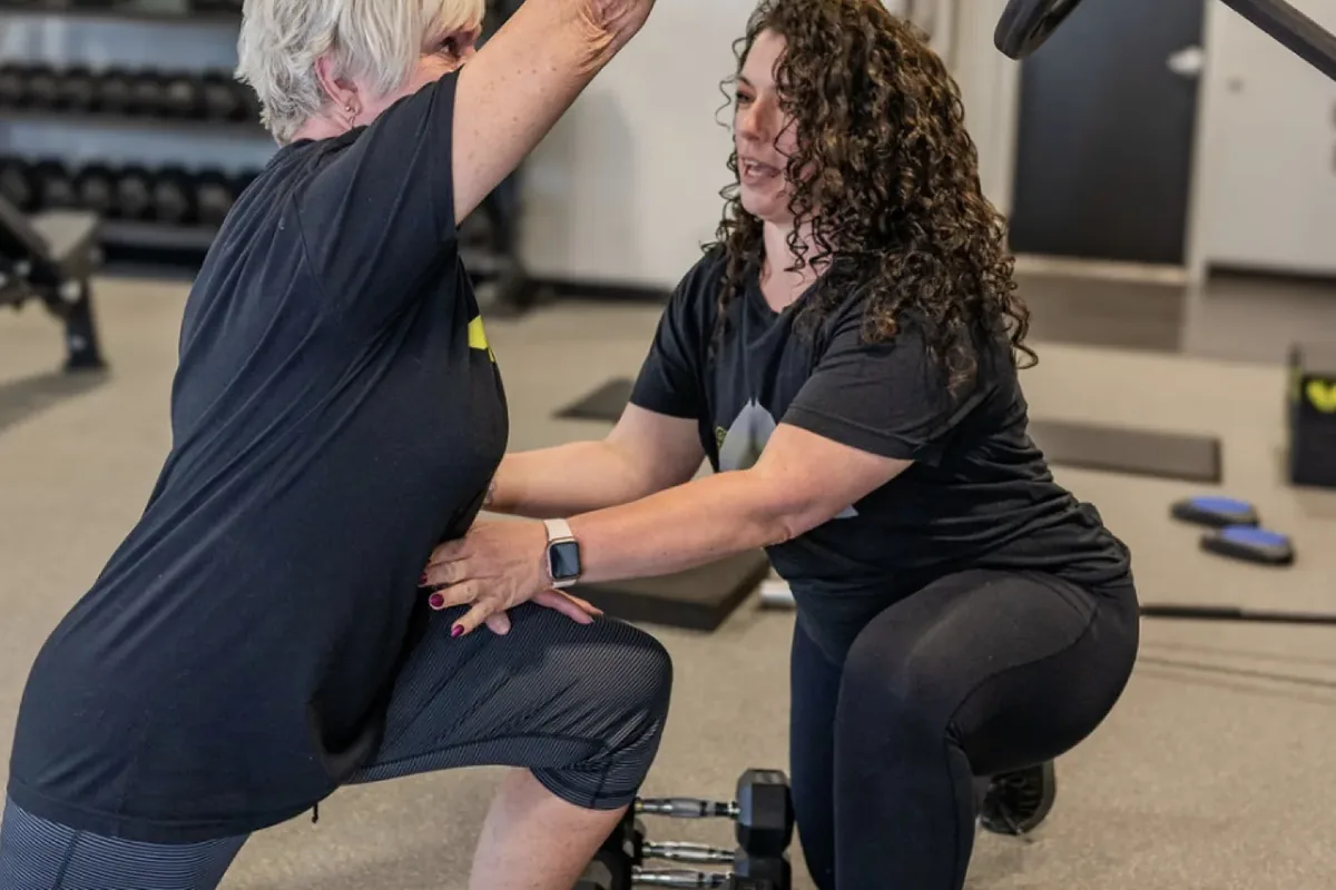 A coach guides an older member through a kneeling single-arm overhead press at Alloy Personal Training Middleton in Middleton, WI.