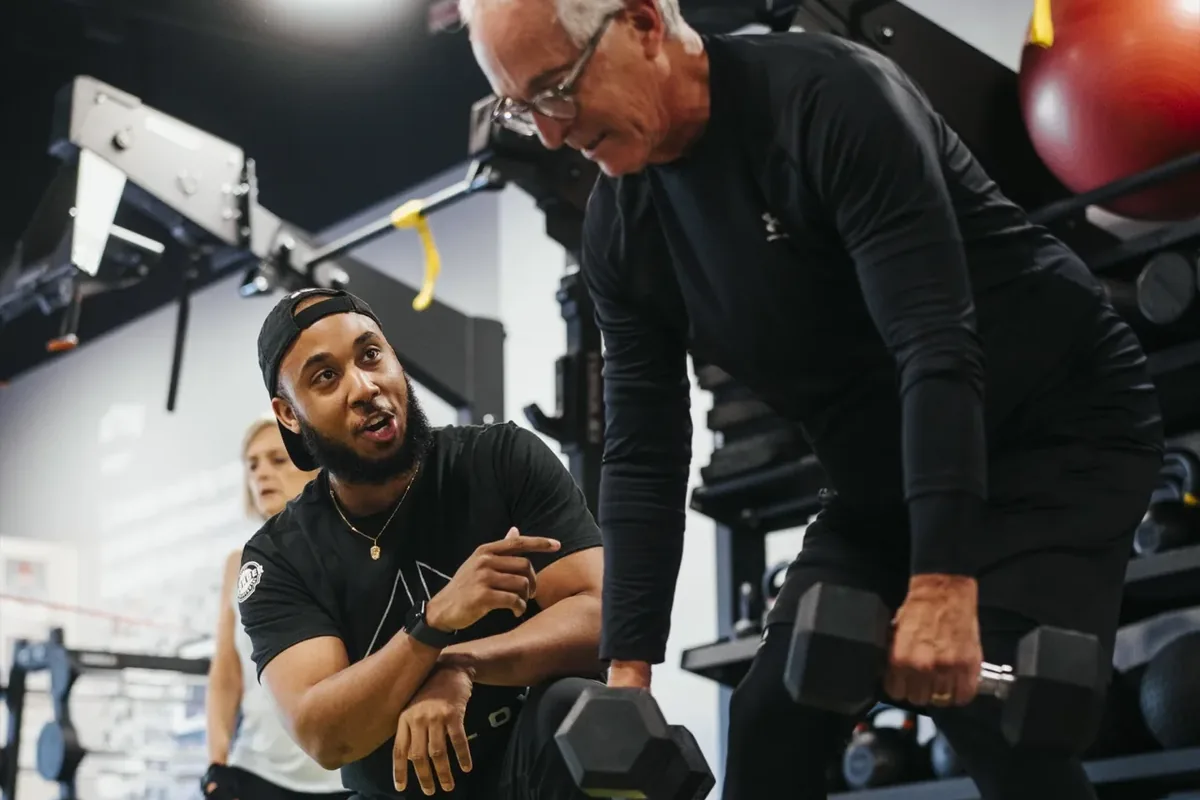 A kneeling coach points out form cues while an older member performs a bent-over dumbbell row at Alloy Personal Training Middleton in Middleton, WI.